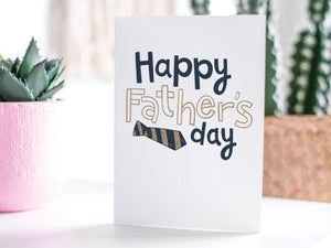 A greeting card featured standing up on a white tabletop with a pink plant pot in the background and some succulents in the pot. There’s a woven basket in the background with a cactus inside. The card features the words “Happy Father’s  Day” with a striped tie on the bottom of the words. 