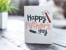Load image into Gallery viewer, A greeting card is featured on a wood coffee table with a green plant in a white planter in the background. The card features the words  “Happy Father’s Day” with an illustrated hammer and screwdriver around the words. 