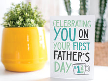 Load image into Gallery viewer, A greeting card is on a table top with a yellow plant pot and a green plant inside. The card features the words &quot;Celebrating you on your first Father&#39;s Day&quot; in modern, simple lettering with a coffee mug with #1 Dad on the mug.