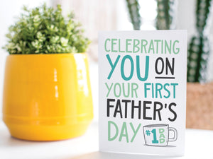 A greeting card is on a table top with a yellow plant pot and a green plant inside. The card features the words "Celebrating you on your first Father's Day" in modern, simple lettering with a coffee mug with #1 Dad on the mug.