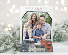 Load image into Gallery viewer, A photo of a one-sided Christmas card showing the front of the card standing up with pine needles behind and blurred white Christmas lights. The photo card features the words “Happy Holidays, The Franklins” at the top with a photo featured in a house shaped frame. Below the photo are illustrated houses covered in snow. 