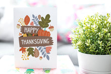 Load image into Gallery viewer, A photo of a card featured on a tabletop next to a white planter filled with a green plant. ​​The card features the words “Happy Thanksgiving” with illustrated leaves, a pumpkin, and acorn.
