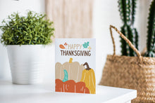Load image into Gallery viewer, A greeting card featured standing up on a white tabletop with a pink plant pot with succulents. There’s a woven basket in the background with a cactus inside. The card features the words “Happy Thanksgiving” with illustrated pumpkins below the words.