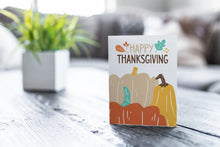 Load image into Gallery viewer, A greeting card featured on a black, wood coffee table. There’s a white planter in the background with a green plant. There’s also a gray sofa in the background with a white pillow. The card features the words &quot;Happy Thanksgiving” with illustrated pumpkins. 