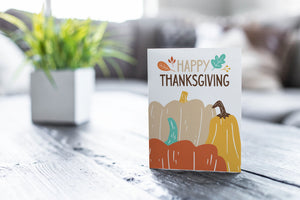 A greeting card featured on a black, wood coffee table. There’s a white planter in the background with a green plant. There’s also a gray sofa in the background with a white pillow. The card features the words "Happy Thanksgiving” with illustrated pumpkins. 