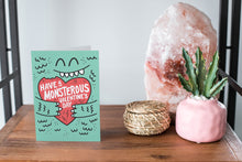 Load image into Gallery viewer, A card on a wood tabletop and on the right side of the card is a woven basket, a pink plant pot with a cactus in it and a pink crystal rock. The card features the words “Have a monstrous Valentine’s Day” with an illustrated monster holding a heart. 