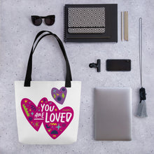 Load image into Gallery viewer, A white tote bag with black handles on a grey tabletop with a laptop and other office supplies. The bag features a design with the words &#39;you are loved&#39; inside two illustrated pink hearts with a smaller, purple heart above. 