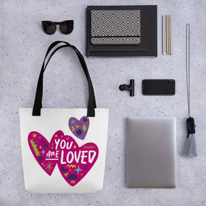 A white tote bag with black handles on a grey tabletop with a laptop and other office supplies. The bag features a design with the words 'you are loved' inside two illustrated pink hearts with a smaller, purple heart above. 