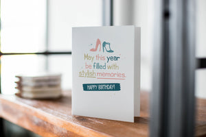 A card on a wood tabletop with an object in the background that is out of focus. The card features the words “May This Year be filled with stylish memories, Happy Birthday” with illustrated heels above the words. 