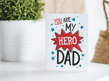 Load image into Gallery viewer, A photo of a card featured on a tabletop next to a white planter filled with a green plant. ​​The card features the words &quot;You are my hero Dad.”