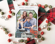 Load image into Gallery viewer, A photo of a one-sided Christmas card showing the front of the card laying on white table top. The card is surrounded by plaid red and green ribbon and red, green and white bells. The photo card features the words “Happy Holidays, The Franklins” at the top with a photo featured in a house shaped frame. Below the photo are illustrated houses covered in snow. 