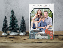 Load image into Gallery viewer, A photo of a one-sided Christmas card showing the front of the card standing up with three small Christmas trees next to it. The photo card features the words “Happy Holidays, The Franklins” at the top with a photo featured in a house shaped frame. Below the photo are illustrated houses covered in snow. 