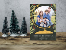 Load image into Gallery viewer, A photo of a one-sided Christmas card showing the front of the card standing up with three small Christmas trees next to it. The photo card features a photo inside an illustrated globe. Around the globe are illustrated leaves. At the bottom of the globe the words “Hope of the World.”