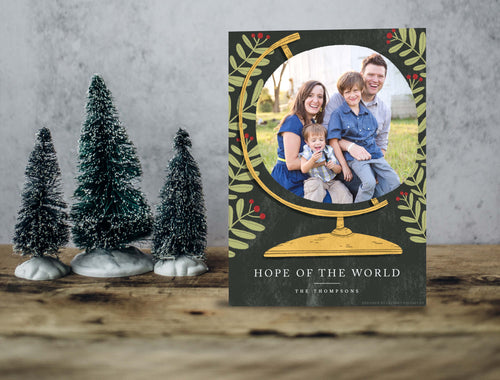 A photo of a one-sided Christmas card showing the front of the card standing up with three small Christmas trees next to it. The photo card features a photo inside an illustrated globe. Around the globe are illustrated leaves. At the bottom of the globe the words “Hope of the World.”