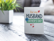 Load image into Gallery viewer, A greeting card is featured on a wood coffee table with a green plant in a white planter in the background. The card features the words “For my Husband on Father&#39;s Day” with small hearts around it. 