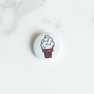 A button pin on a white tabletop. The pin features an illustration of an ice cream cone.