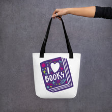 Load image into Gallery viewer, A person holding a white tote bag with black handles. The tote bag has a design with the words &#39;I love books&#39; with the word love shown as a heart. The words are inside an illustrated book. 