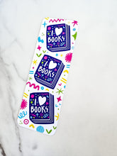 Load image into Gallery viewer, A bookmark on a tabletop. The bookmark features the words &quot;i love books&quot; with love shown shaped as a heart. The words are placed inside an illustrated book. 