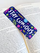 Load image into Gallery viewer, A bookmark placed inside a book. The bookmark has the words &quot;stay curious&quot; with fun, sketchy illustrations. 