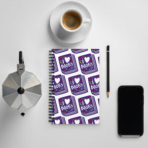 A spiral notebook on a tabletop with a coffee mug and phone around it. The white spiral notebook has black spiral binding. The pattern design features the words "I love books" inside an illustrated book with the word love as a heart. 