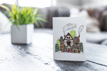 Load image into Gallery viewer, A greeting card featured on a black, wood coffee table. There’s a white planter in the background with a green plant. There’s also a gray sofa in the background with a white pillow. The card features an illustrated English village covered in snow with pine trees and smoke coming from the chimneys.