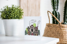 Load image into Gallery viewer, A greeting card is featured on a white tabletop with a white planter in the background with a green plant. There’s a woven basket in the background with a cactus inside. The card features an illustrated English village covered in snow with pine trees and smoke coming from the chimneys. 