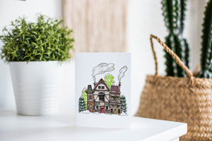 A greeting card is featured on a white tabletop with a white planter in the background with a green plant. There’s a woven basket in the background with a cactus inside. The card features an illustrated English village covered in snow with pine trees and smoke coming from the chimneys. 