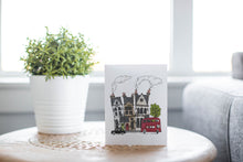 Load image into Gallery viewer, A greeting card laying on a wooden table with some cut wood details. The card features a design with illustrated London houses, a black taxi cab and a red double decker bus. 