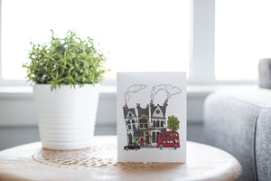 A greeting card laying on a wooden table with some cut wood details. The card features a design with illustrated London houses, a black taxi cab and a red double decker bus. 