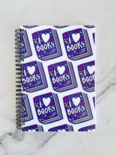 Load image into Gallery viewer, A spiral notebook is on a tabletop. The white spiral notebook has black spiral binding. The pattern design features the words &quot;I love books&quot; inside an illustrated book with the word love as a heart. 