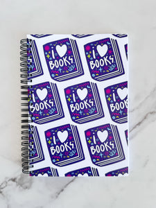 A spiral notebook is on a tabletop. The white spiral notebook has black spiral binding. The pattern design features the words "I love books" inside an illustrated book with the word love as a heart. 