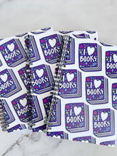 Load image into Gallery viewer, A closeup of three spiral notebooks are on a tabletop. The white spiral notebooks have black spiral binding. The pattern design features the words &quot;I love books&quot; inside an illustrated book with the word love as a heart. 