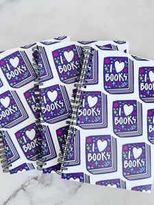 A closeup of three spiral notebooks are on a tabletop. The white spiral notebooks have black spiral binding. The pattern design features the words "I love books" inside an illustrated book with the word love as a heart. 