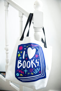 A white tote bag with black handles hanging on a staircase. The tote bag has a design with the words 'I love books' with the word love shown as a heart. The words are inside an illustrated book. 