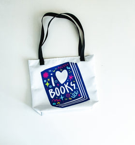 A white tote bag with black handles laying on a white tabel. The tote bag has a design with the words 'I love books' with the word love shown as a heart. The words are inside an illustrated book. 