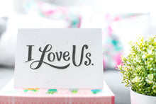 Load image into Gallery viewer, A greeting card is on a table top with a gift in pink wrapping paper. Next to the gift is a white plant pot with a green plant. The card features the words “I Love Us.”