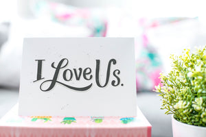 A greeting card is on a table top with a gift in pink wrapping paper. Next to the gift is a white plant pot with a green plant. The card features the words “I Love Us.”