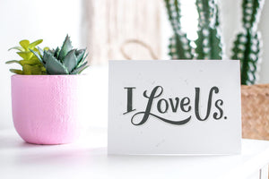 A greeting card featured standing up on a white tabletop with a pink plant pot in the background and some succulents in the pot. There’s a woven basket in the background with a cactus inside. The card features the words “I Love Us.”