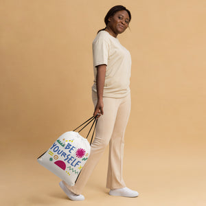 A woman carrying a white drawstring bag. The drawstring bag has the words "Be Yourself" in blue with green illustrated sketchy pieces around the words. 