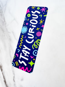 A bookmark placed on a tabletop. The bookmark has the words "stay curious" with fun, sketchy illustrations. 