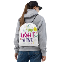 Load image into Gallery viewer, A woman holding a white drawstring bag on her back. She&#39;s wearing a grey hoodie and a black hat. The drawstring bag has the words &quot;Let Your Light Shine&quot; with sketchy illustrations around the words.