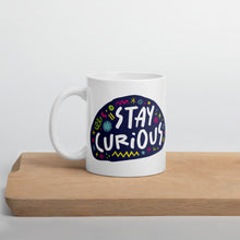 Load image into Gallery viewer, A white mug on a wood board. The mug features a design with the words &#39;stay curious&#39; in an illustrated oval shape with sketchy elements around it. 