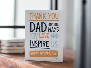 A card on a wood tabletop with an object in the background that is out of focus. The card features the words "Thank You Dad for the ways you love and inspire us. Happy Father's Day.” 