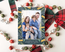 Load image into Gallery viewer, A photo of a one-sided Christmas card showing the front of the card laying on white table top. The card is surrounded by plaid red and green ribbon and red, green and white bells. The photo card features one photo with a border of illustrated gold crowns, and green and white illustrated leaves. The bottom of the card reads “Celebrate the Prince of Peace, The Thompsons.”