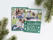 Load image into Gallery viewer, A photo of a Christmas card showing the front and back of the card laying on a white surface. Around the two sides of the card are pine needles. The front of the card features a photo on the top and on the bottom is in green with words in white “Hallelujah Jesus is Here” with white stars around it. The back of the card features two photos. 