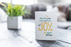 A greeting card is featured on a wood coffee table with a green plant in a white planter in the background. The card features the words “May This Year Bring You Lots of Joy Happy Birthday.”