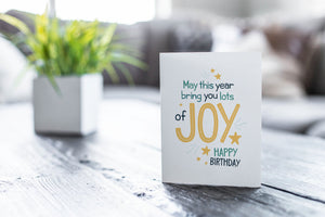 A greeting card featured on a black, wood coffee table. There’s a white planter in the background with a green plant. There’s also a gray sofa in the background with a white pillow. The card features the words “May This Year Bring You Lots of Joy, Happy Birthday.”