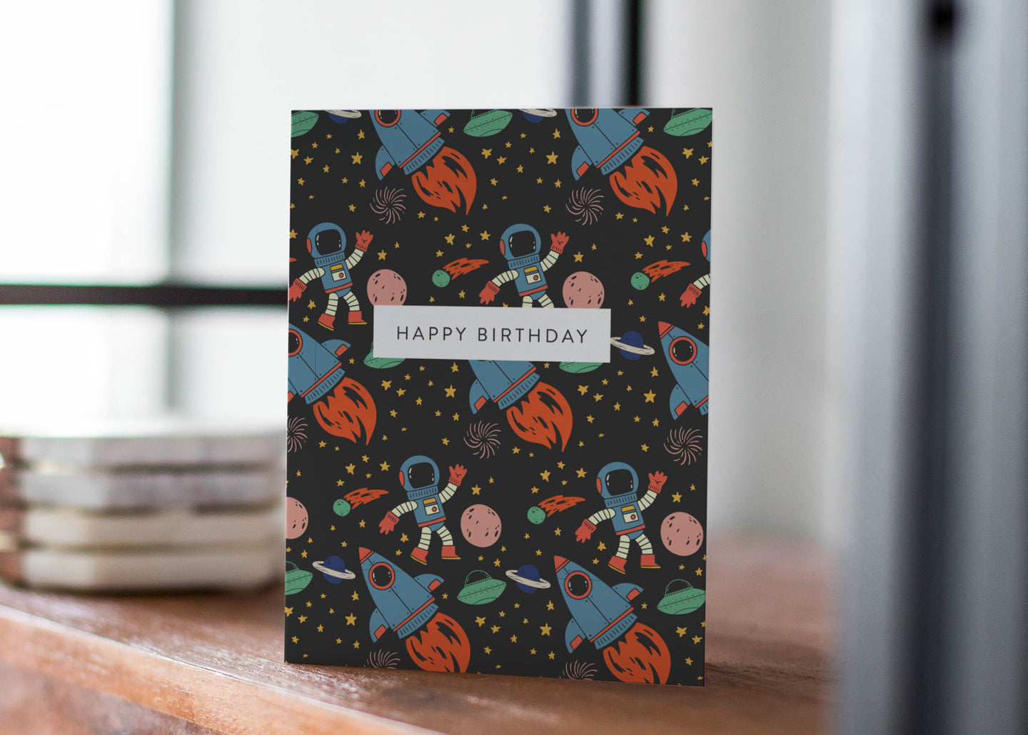 A greeting card standing up on a table with coasters in the background. The greeting card features the words 'happy birthday' with illustrated space items around it. The space items include an astronaut, space rockets, planets and stars. 