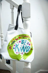 A white tote bag with black handles hanging on a staircase. The bag features a design with the words 'be kind' with illustrated hugging hands around the words. 