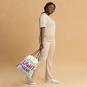 A woman walking while holding a white drawstring bag. The drawstring bag has the words "Let Your Light Shine" with sketchy illustrations around the words.